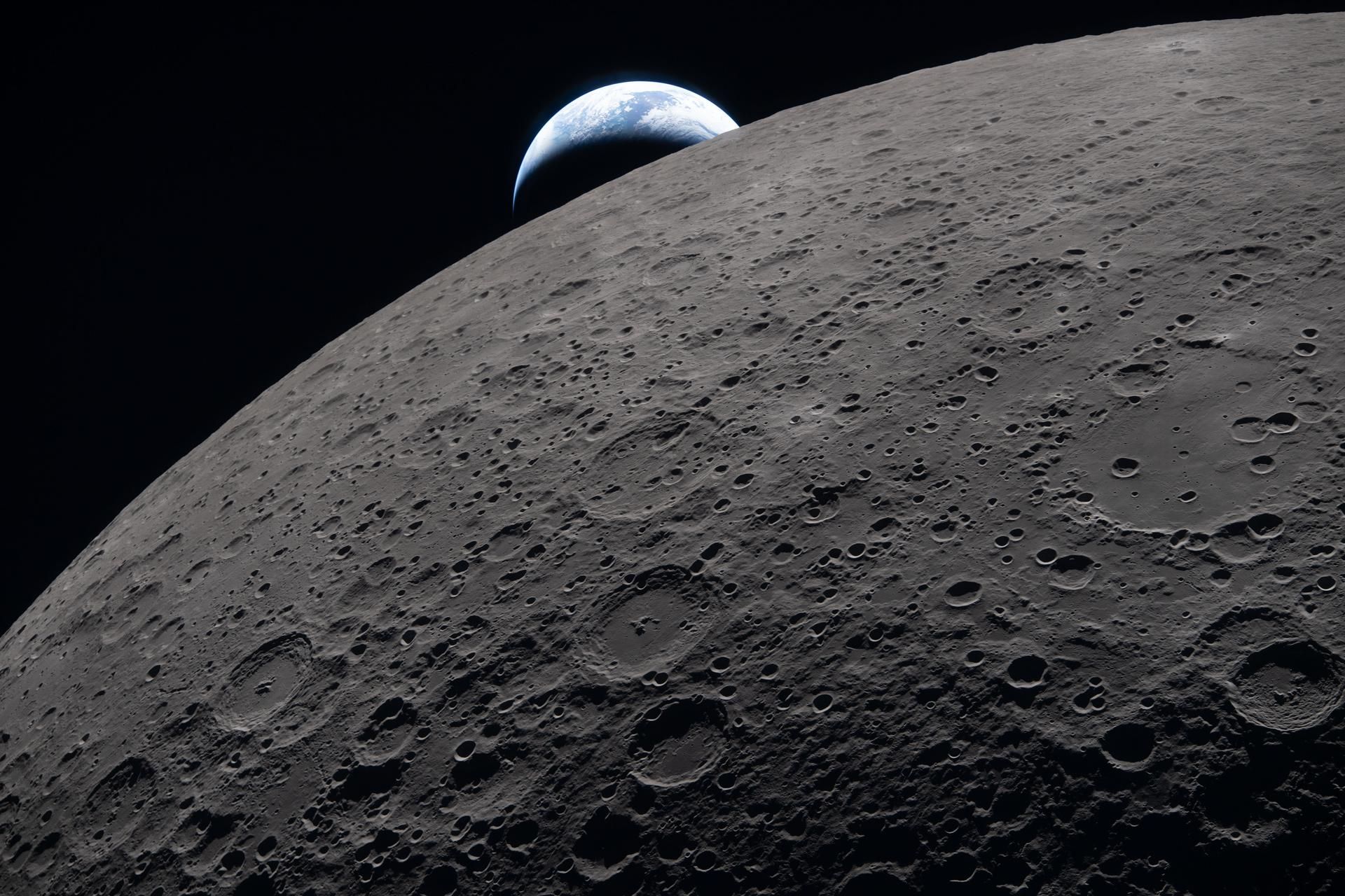 The lunar surface fills the frame in sharp detail, as seen during the Artemis II lunar flyby, while a distant Earth sets in the background. This image was captured at 6:41 p.m. EDT, on April 6, 2026, just three minutes before the Orion spacecraft and its crew went behind the Moon and lost contact with Earth for 40 minutes before emerging on the other side. In this image, the dark portion of Earth is experiencing nighttime, while on its day side, swirling clouds are visible over the Australia and Oceania region. In the foreground, Ohm crater shows terraced edges and a relatively flat floor marked by central peaks — formed when the surface rebounded upward during the impact that created the crater. 